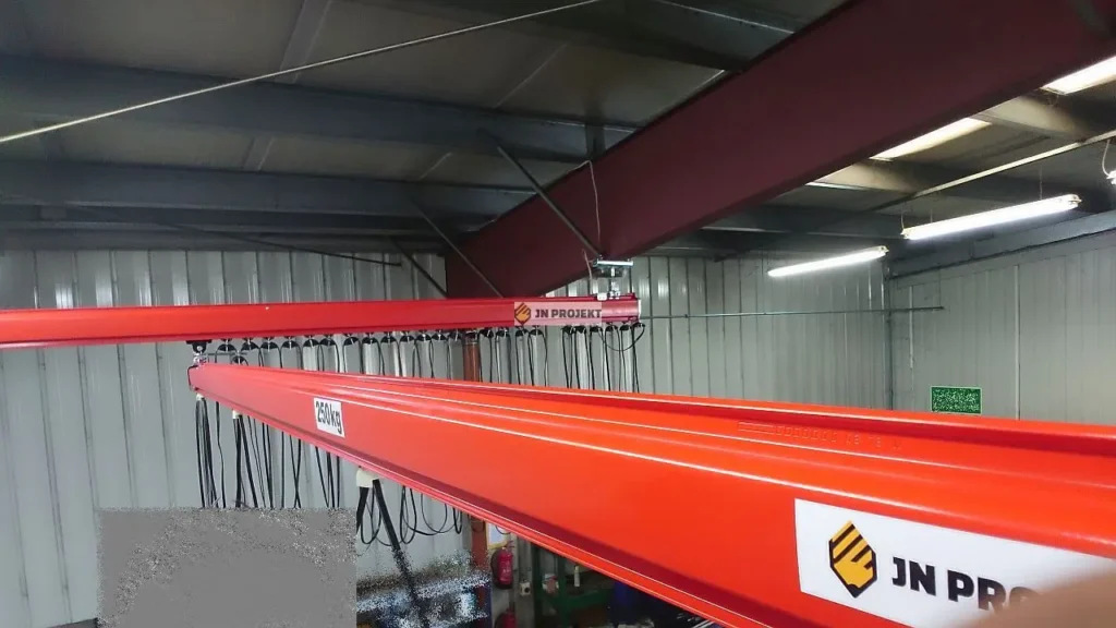 Czerwona suwnica modułowa z wciągnikiem łańcuchowym podwieszona do konstrukcji dachu hali